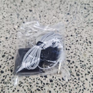May include: A small, black electronic device with a white cord and a gold-colored plug, packaged in a clear plastic bag. The cord is coiled and neatly arranged. The background is a speckled, gray surface.