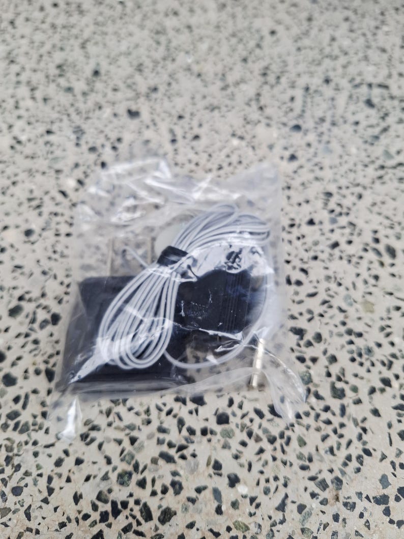May include: A small, black electronic device with a white cord and a gold-colored plug, packaged in a clear plastic bag. The cord is coiled and neatly arranged. The background is a speckled, gray surface.