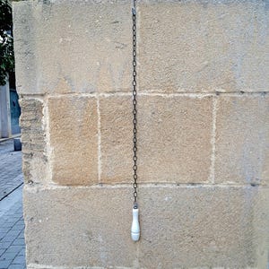May include: A white ceramic pull chain hanging from a black chain attached to a hook on a stone wall.