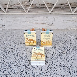 Puede incluir: Tres encendedores de cigarrillos en miniatura con forma de paquetes de cigarrillos Camel. Los encendedores son plateados con un dise&ntilde;o amarillo y azul. Los encendedores presentan el logotipo de Camel y el texto "Camel Filters Turkish & American Blend Cigarettes".