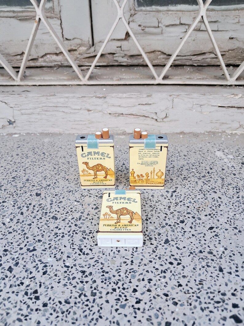 Puede incluir: Tres encendedores de cigarrillos en miniatura con forma de paquetes de cigarrillos Camel. Los encendedores son plateados con un dise&ntilde;o amarillo y azul. Los encendedores presentan el logotipo de Camel y el texto "Camel Filters Turkish & American Blend Cigarettes".