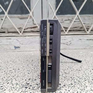 May include: A gray and beige portable radio with an extendable antenna. The radio has a volume control, tuning dial, and various function buttons. The side view shows the radio's controls and a black cord.