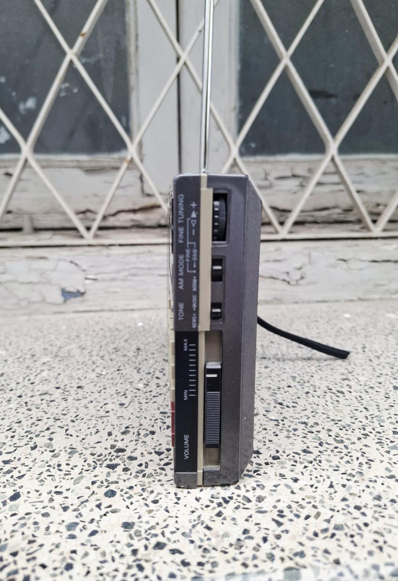 May include: A gray and beige portable radio with an extendable antenna. The radio has a volume control, tuning dial, and various function buttons. The side view shows the radio's controls and a black cord.