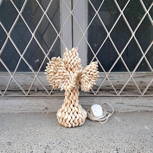 May include: A decorative cross made from small, pale shells. It sits on a grey surface, with a white electrical plug and cable to the right. The background shows a window with a diamond-patterned metal grille.