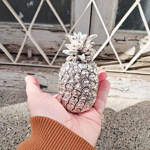 May include: A silver-colored pineapple sculpture with detailed textures, held in a hand. The pineapple's crown and body are intricately designed, reflecting light. The sculpture is a decorative item, possibly for home decor or as a unique gift.