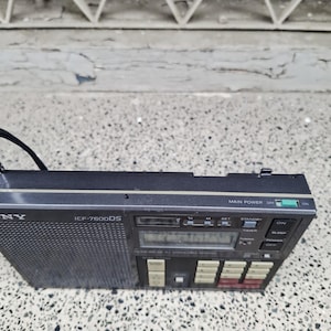 May include: A black Sony ICF-7600DS portable radio with a mesh speaker cover and a digital display. The radio has several buttons and a tuning dial. The main power switch is set to off.