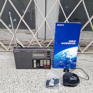 May include: A black Sony ICF-7600DS radio with an antenna, a blue "WAVE HANDBOOK", a black AM-91 cassette player, and a set of white earbuds. The radio has a digital display and several buttons. The items are on a speckled surface.