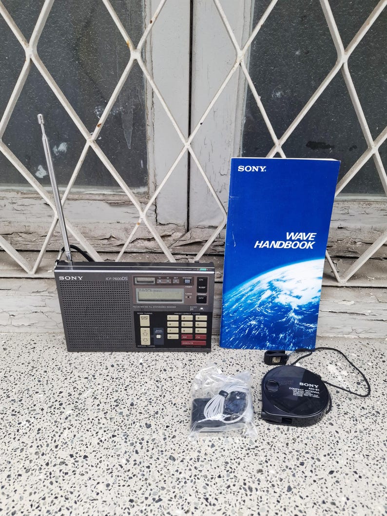 May include: A black Sony ICF-7600DS radio with an antenna, a blue "WAVE HANDBOOK", a black AM-91 cassette player, and a set of white earbuds. The radio has a digital display and several buttons. The items are on a speckled surface.