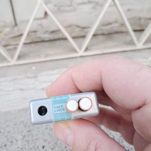 Puede incluir: Un dispositivo rectangular de color plateado con dos botones blancos y una peque&ntilde;a lente negra. El dispositivo tiene una etiqueta azul y blanca que dice "BACK LITES".