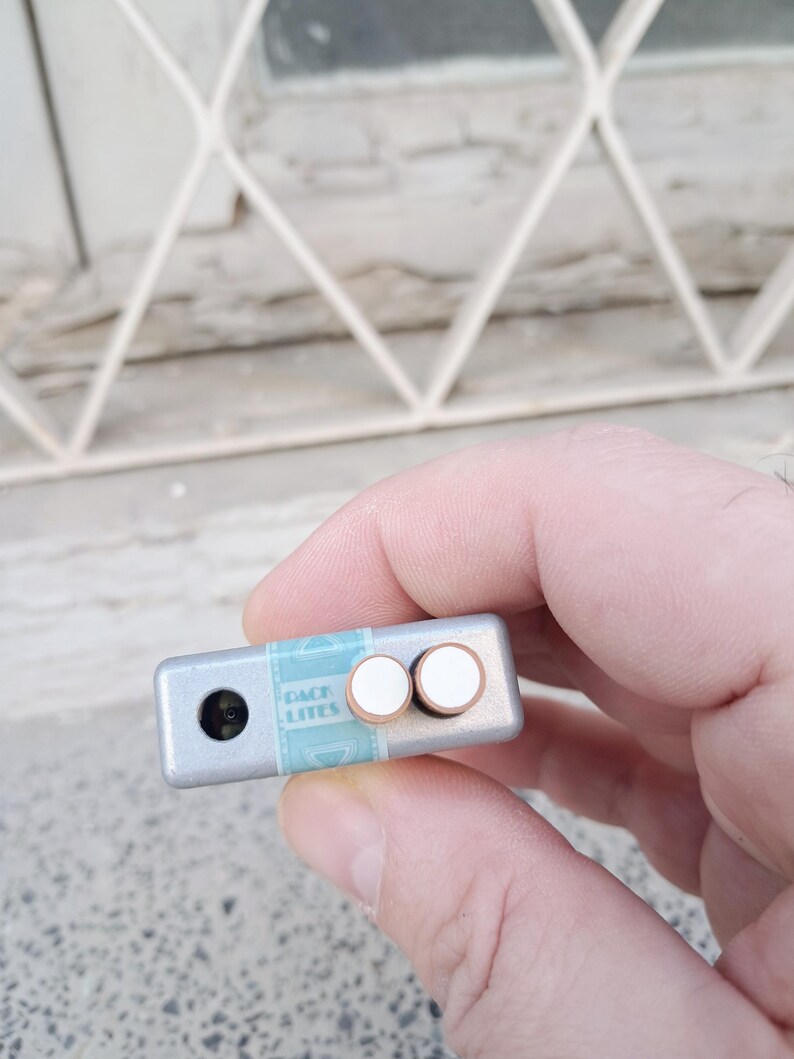 Puede incluir: Un dispositivo rectangular de color plateado con dos botones blancos y una peque&ntilde;a lente negra. El dispositivo tiene una etiqueta azul y blanca que dice "BACK LITES".