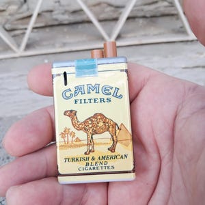 Puede incluir: Una r&eacute;plica en miniatura de un paquete de cigarrillos Camel, con una ilustraci&oacute;n de camello y el texto "Camel Filters Turkish & American Blend Cigarettes".