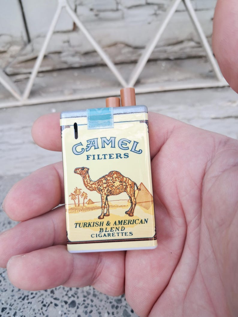 Puede incluir: Una r&eacute;plica en miniatura de un paquete de cigarrillos Camel, con una ilustraci&oacute;n de camello y el texto "Camel Filters Turkish & American Blend Cigarettes".
