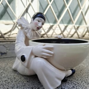 Puede incluir: Una figura de cerámica de un payaso pierrot sentado con cuello de volantes y traje blanco, con botones negros y un gorro negro. El payaso sostiene un cuenco grande, liso y de color crema con un interior oscuro, formando una pieza decorativa.