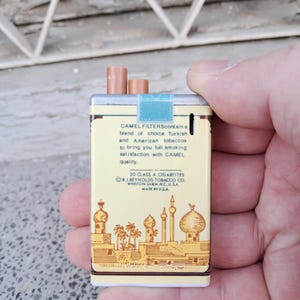 Puede incluir: Un paquete vintage de cigarrillos Camel Filter con fondo amarillo y una representaci&oacute;n de una ciudad de Oriente Medio. El texto en el paquete dice "Camel Filters contain a blend of choice Turkish and American tobaccos to bring you full smoking satisfaction with Camel quality. 20 Class A Cigarettes R.J. Reynolds Tobacco Co. Winston-Salem, N.C. U.S.A. Made in U.S.A."