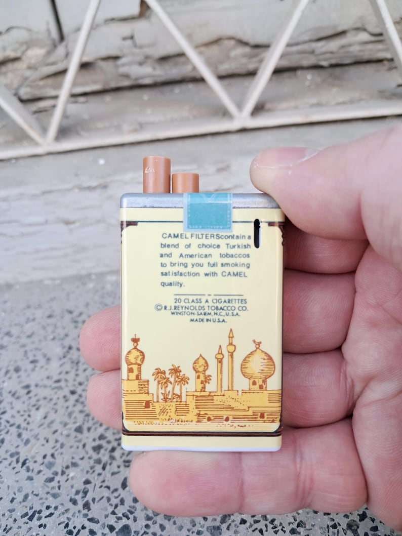 Puede incluir: Un paquete vintage de cigarrillos Camel Filter con fondo amarillo y una representaci&oacute;n de una ciudad de Oriente Medio. El texto en el paquete dice "Camel Filters contain a blend of choice Turkish and American tobaccos to bring you full smoking satisfaction with Camel quality. 20 Class A Cigarettes R.J. Reynolds Tobacco Co. Winston-Salem, N.C. U.S.A. Made in U.S.A."