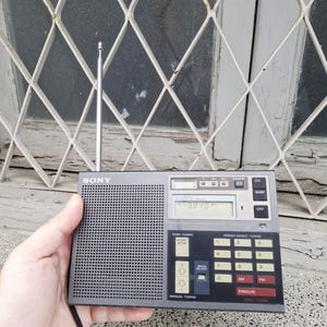 May include: A gray Sony portable radio with a retractable antenna. The radio has a digital display showing "9840" and features buttons for scan tuning, preset tuning, and manual tuning. The radio is held in a hand.