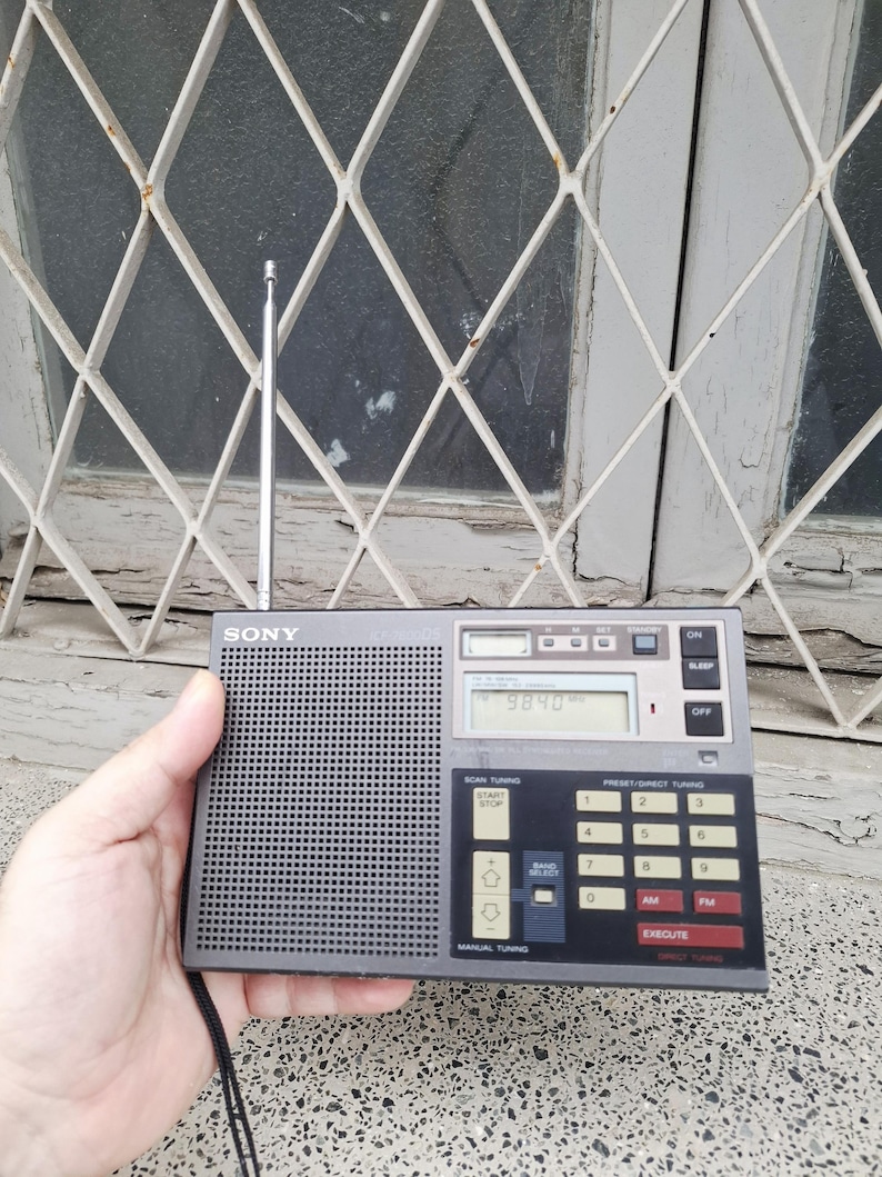 May include: A gray Sony portable radio with a retractable antenna. The radio has a digital display showing "9840" and features buttons for scan tuning, preset tuning, and manual tuning. The radio is held in a hand.