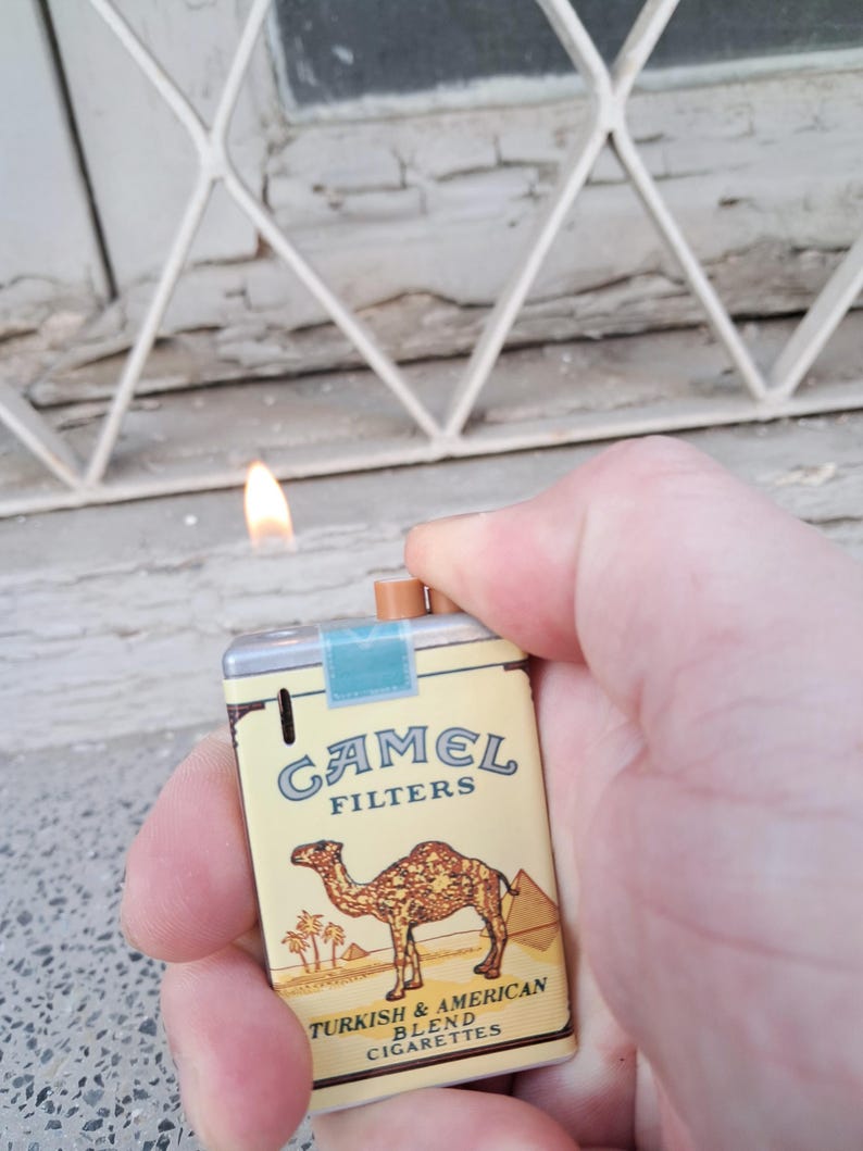 May include: A miniature lighter shaped like a pack of Camel cigarettes. The lighter is yellow and blue with a camel illustration and the text "Camel Filters Turkish & American Blend Cigarettes".