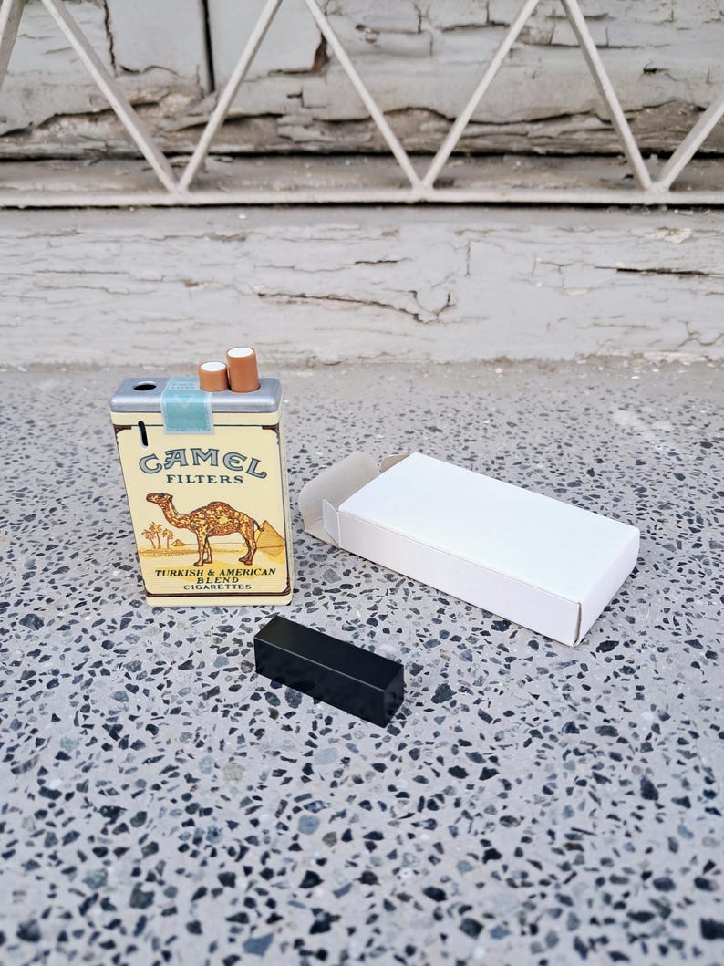 May include: A vintage-style Camel cigarette pack lighter featuring a camel illustration and the text "Camel Filters Turkish & American Blend Cigarettes". The lighter is silver with a blue and yellow design. It is sitting on a white surface with a black lighter next to it.