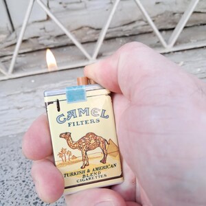 Puede incluir: Una r&eacute;plica en miniatura de un paquete de cigarrillos Camel. El paquete es amarillo con una ilustraci&oacute;n de camello marr&oacute;n y el texto "Camel Filters Turkish & American Blend Cigarettes".