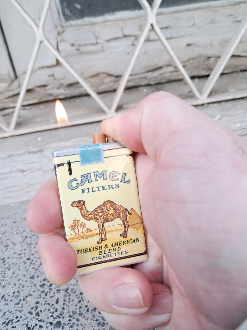 Puede incluir: Una r&eacute;plica en miniatura de un paquete de cigarrillos Camel. El paquete es amarillo con una ilustraci&oacute;n de camello marr&oacute;n y el texto "Camel Filters Turkish & American Blend Cigarettes".