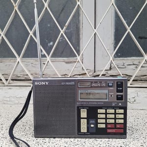 May include: A gray Sony ICF-7600DS portable radio with a retractable antenna. The radio has a digital display showing 98.40 and various buttons for tuning and functions. A black strap is attached to the side.