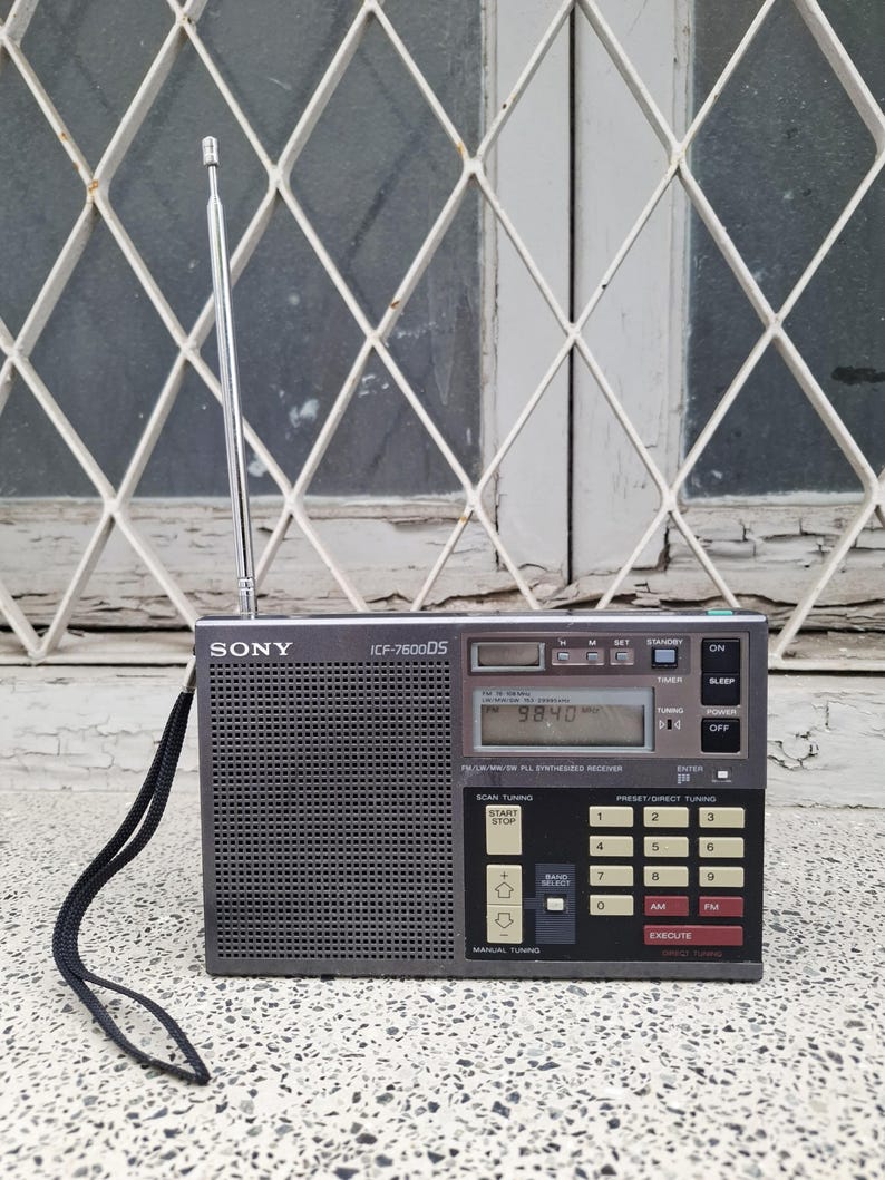 May include: A gray Sony ICF-7600DS portable radio with a retractable antenna. The radio has a digital display showing 98.40 and various buttons for tuning and functions. A black strap is attached to the side.