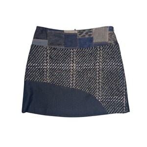 Op de afbeelding: Een patchwork minirok met een mix van stoffen en patronen. De rok combineert denim, ruitjes en getextureerde materialen in tinten blauw, grijs en neutrale tinten. Het ontwerp bevat een gebogen paneel aan de onderkant.