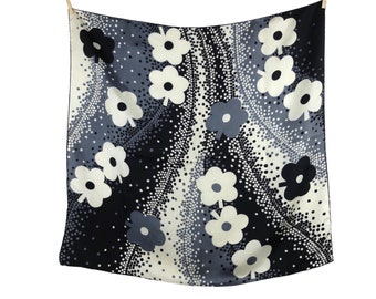 May include: A black and white silk scarf with a floral pattern. The flowers are white with black centres and are scattered across the scarf. The background is a dark grey with white dots.