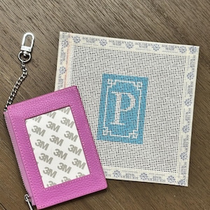 May include: A bright pink pebbled leather card holder with a silver chain and lobster clasp, featuring a clear window with 3M branding. Beside it is a square needlepoint canvas with a blue border and a large white letter P in the center.