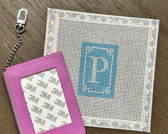 May include: A bright pink pebbled leather card holder with a silver chain and lobster clasp, featuring a clear window with 3M branding. Beside it is a square needlepoint canvas with a blue border and a large white letter P in the center.