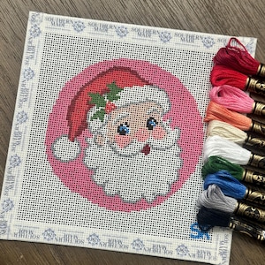 May include: A square needlepoint canvas featuring a Santa Claus head design on a pink circular background, with a white border printed with the text Southern Made. A stack of ten colorful embroidery floss skeins is positioned on the right side.