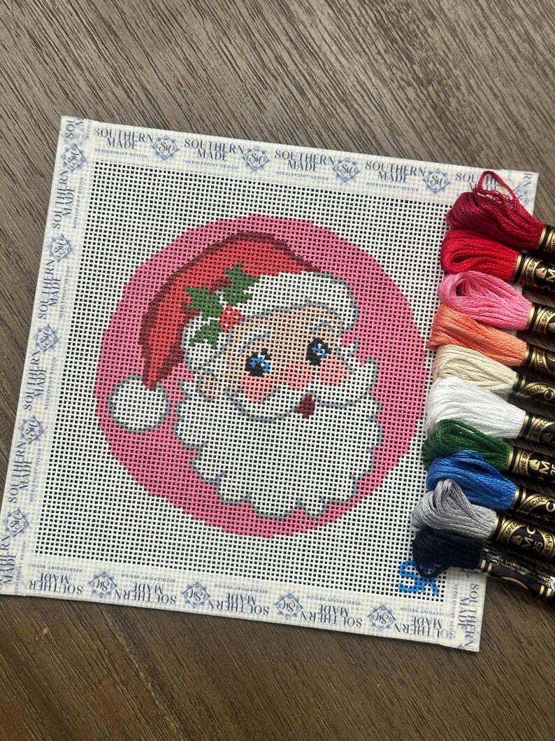 May include: A square needlepoint canvas featuring a Santa Claus head design on a pink circular background, with a white border printed with the text Southern Made. A stack of ten colorful embroidery floss skeins is positioned on the right side.