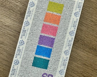 May include: A needlepoint canvas with a gradient of colors: yellow, orange, pink, purple, blue, and teal. The canvas is labeled "SOUTHERN MADE" around the edges and has "SR" in the bottom corner.