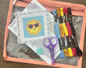 May include: A needlepoint kit in a peach zippered pouch containing a square canvas with a yellow sun wearing pink sunglasses, instructional papers, a pair of purple scissors, a small light blue flower charm, and six skeins of embroidery floss.