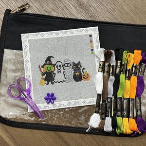 May include: A needlepoint canvas featuring a witch, ghost, mummy, and black cat with a pumpkin, accompanied by purple scissors, a purple flower-shaped needle minder, and eight skeins of embroidery floss in white, brown, gray, black, green, yellow, orange, and purple.