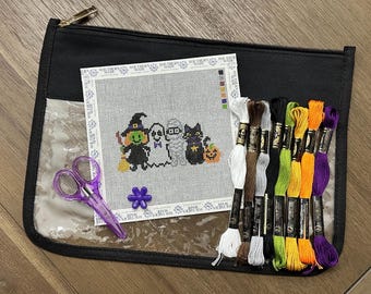 May include: A needlepoint canvas featuring a witch, ghost, mummy, and black cat with a pumpkin, accompanied by purple scissors, a purple flower-shaped needle minder, and eight skeins of embroidery floss in white, brown, gray, black, green, yellow, orange, and purple.