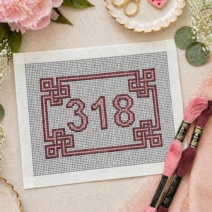 May include: A cross-stitch project with the number "318" in a pink hue, framed by a decorative border. Two spools of pink and burgundy embroidery floss are in the foreground. The background is a light, neutral color.