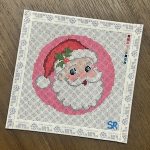 May include: A square needlepoint canvas featuring a circular pink background with a Santa Claus face wearing a red hat with holly leaves. The border is printed with the text Southern Made repeatedly, and the initials SR are in the bottom right corner.