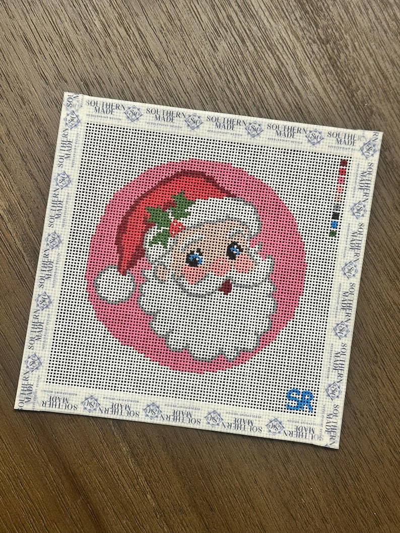 May include: A square needlepoint canvas featuring a circular pink background with a Santa Claus face wearing a red hat with holly leaves. The border is printed with the text Southern Made repeatedly, and the initials SR are in the bottom right corner.