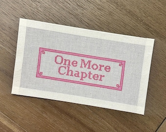 May include: A rectangular needlepoint canvas with the text "One More Chapter" in pink within a pink rectangular border. The canvas is white with a grid pattern, and the background is a wooden surface.