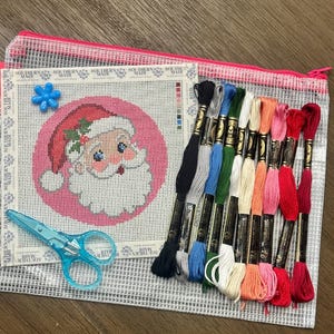 May include: A needlepoint canvas featuring a Santa Claus design on a pink circular background, accompanied by eleven skeins of embroidery floss in various colors, a blue plastic needle threader, and light blue scissors, all inside a mesh pouch.