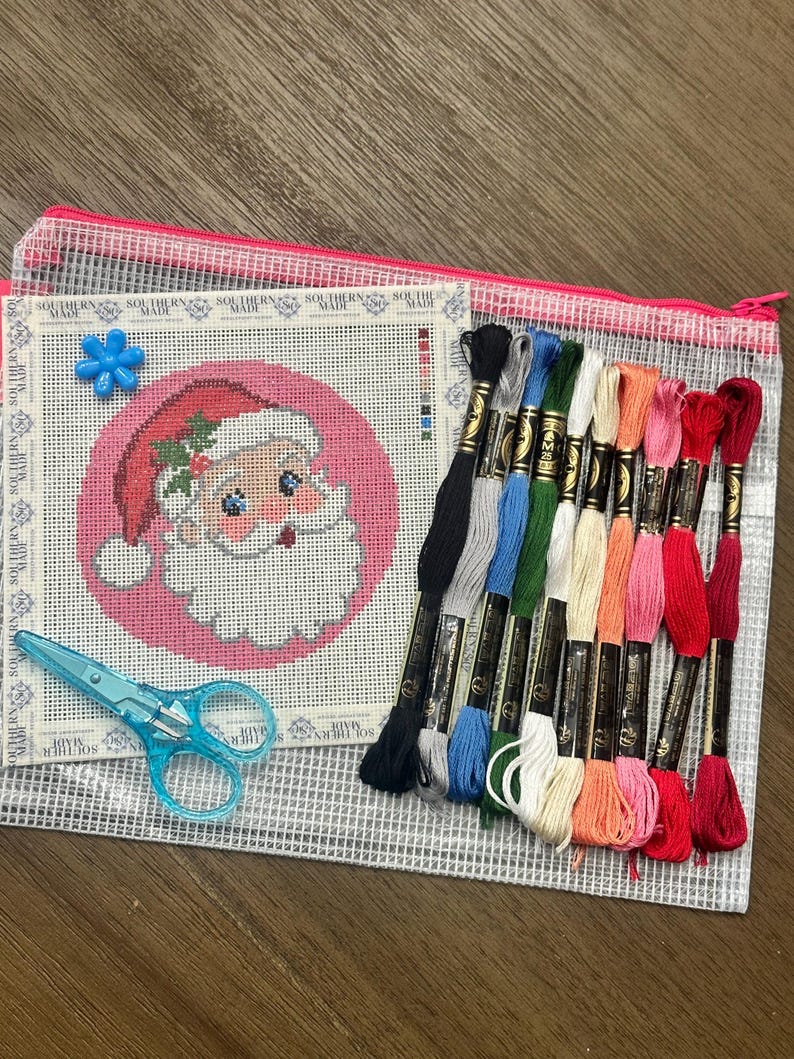 May include: A needlepoint canvas featuring a Santa Claus design on a pink circular background, accompanied by eleven skeins of embroidery floss in various colors, a blue plastic needle threader, and light blue scissors, all inside a mesh pouch.