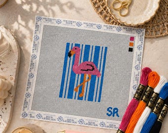 May include: A needlepoint canvas featuring a pink flamingo against blue stripes. The canvas is surrounded by a white border with the text "SOUTHERN MADE". A pink pen with the text "STITCH & UNWIND" and embroidery floss in various colors are also present.