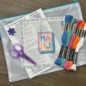 May include: A needlepoint kit with a clear zippered pouch, scissors, and embroidery floss. The kit includes a canvas with the letters "MA HJ" stitched in colorful thread, a step-by-step guide, and a needlepoint pattern.