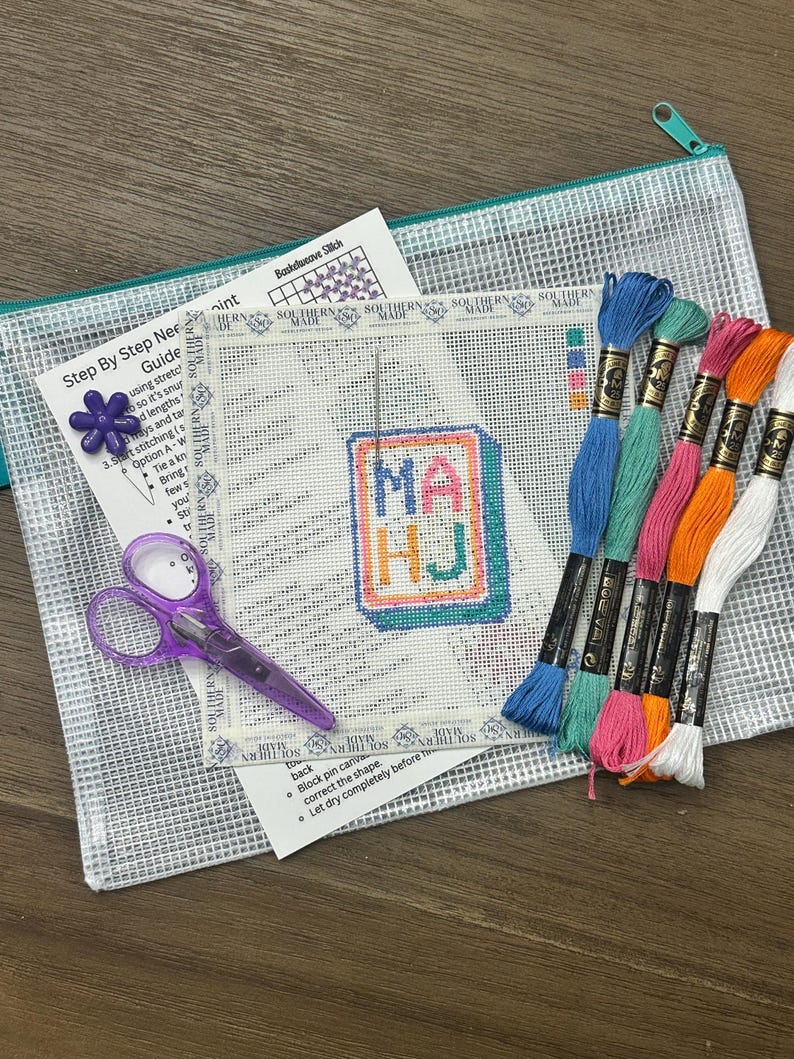 May include: A needlepoint kit with a clear zippered pouch, scissors, and embroidery floss. The kit includes a canvas with the letters "MA HJ" stitched in colorful thread, a step-by-step guide, and a needlepoint pattern.