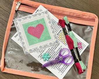 May include: A needlepoint kit in a clear peach-colored zippered pouch containing a square canvas with a pink heart on a green background, two skeins of embroidery floss in pink and light green, a small flower-shaped bead, and printed instructions.