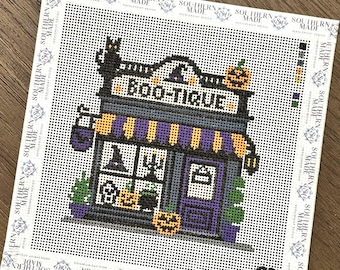 May include: A needlepoint canvas featuring a storefront with the text BOO-TIQUE, a black cat, pumpkins, and a witch hat. The design includes purple and yellow striped awnings, a purple door, and a color palette of black, purple, orange, and green.