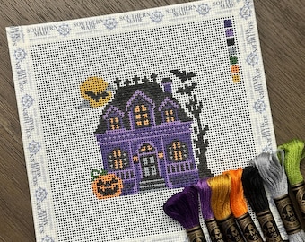 May include: A square needlepoint canvas featuring a purple haunted house with a black roof, a yellow moon, bats, and a jack-o'-lantern. The border includes the text Southern Made. Seven skeins of embroidery floss in purple, gold, orange, black, and gray are included.