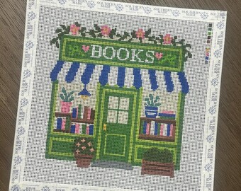 May include: A square needlepoint canvas featuring a storefront with a green frame, blue and white striped awning, and the word BOOKS in white text. The design includes pink flowers, potted plants, and shelves filled with colorful books.